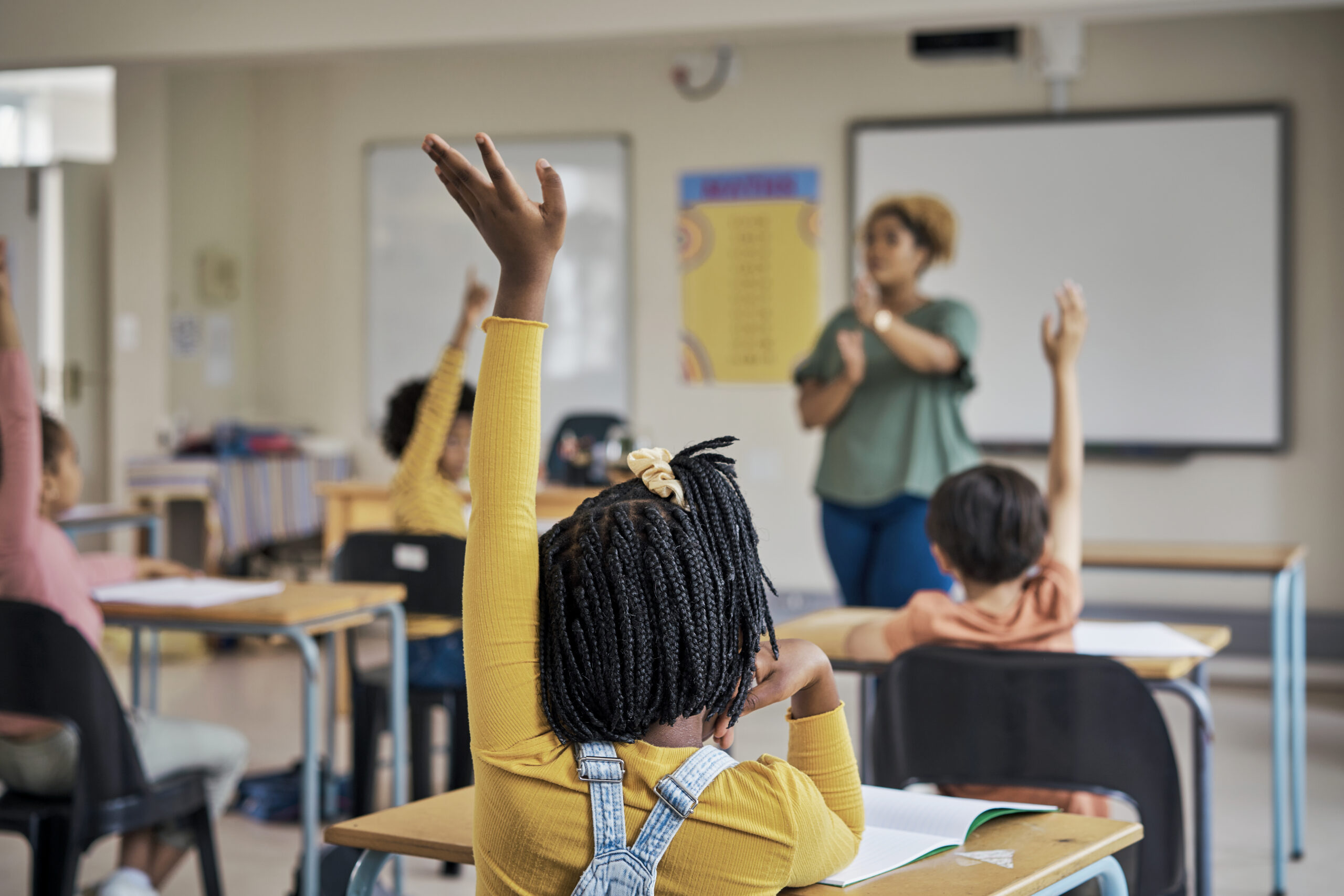 Multicultural elementary classroom showing students seated at their desks, raising hands to answer teacher's math question.