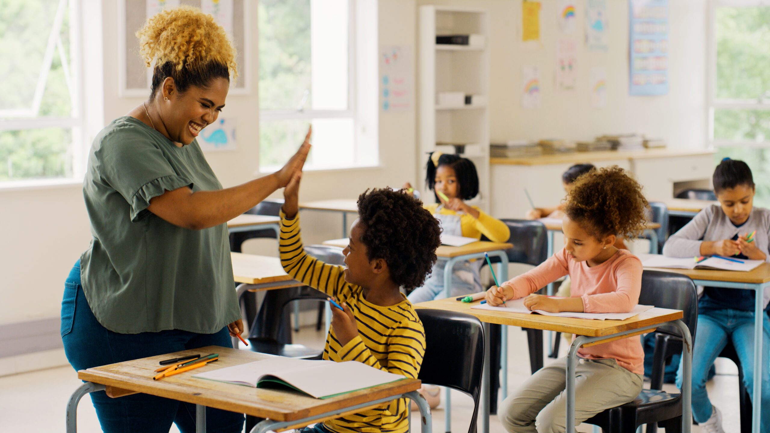 A black female teacher giving a black elementary student a high-five in a classroom with other students.