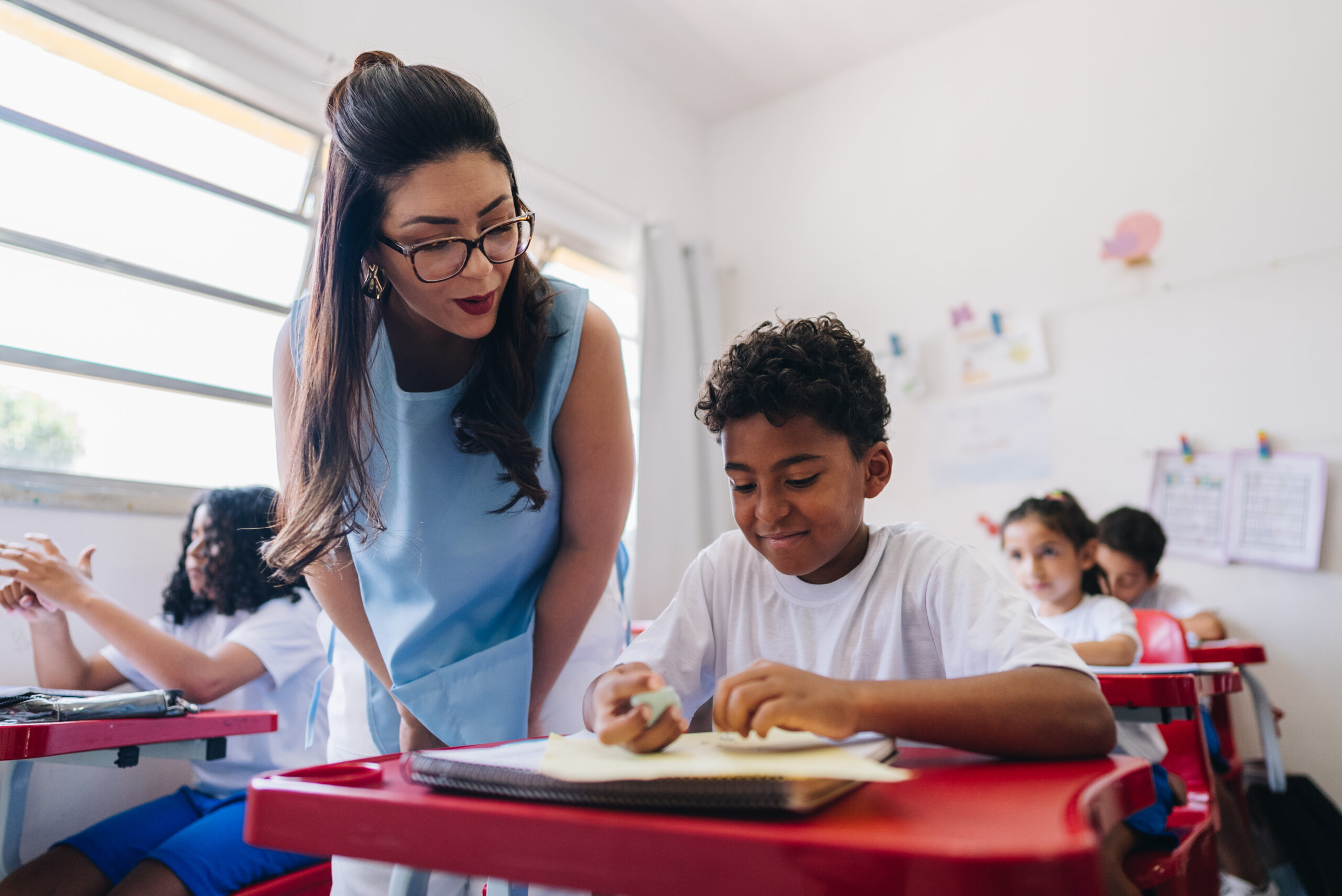 Hispanic female teacher in classroom walking up to student desk during math lesson.