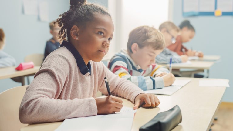 Image of elementary school students writing at desk, with young Black girl in the front looking curious.
