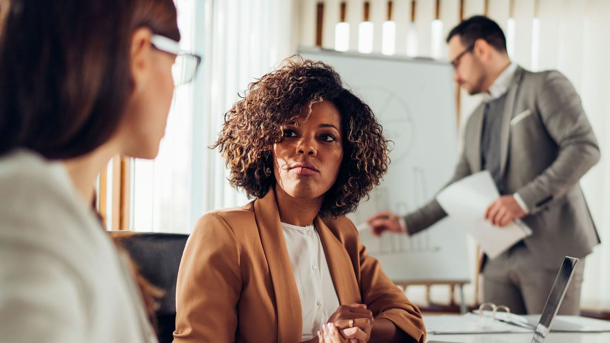 Black Woman School Leader Looking at Another Woman With Male in Background Writing on a Whiteboard