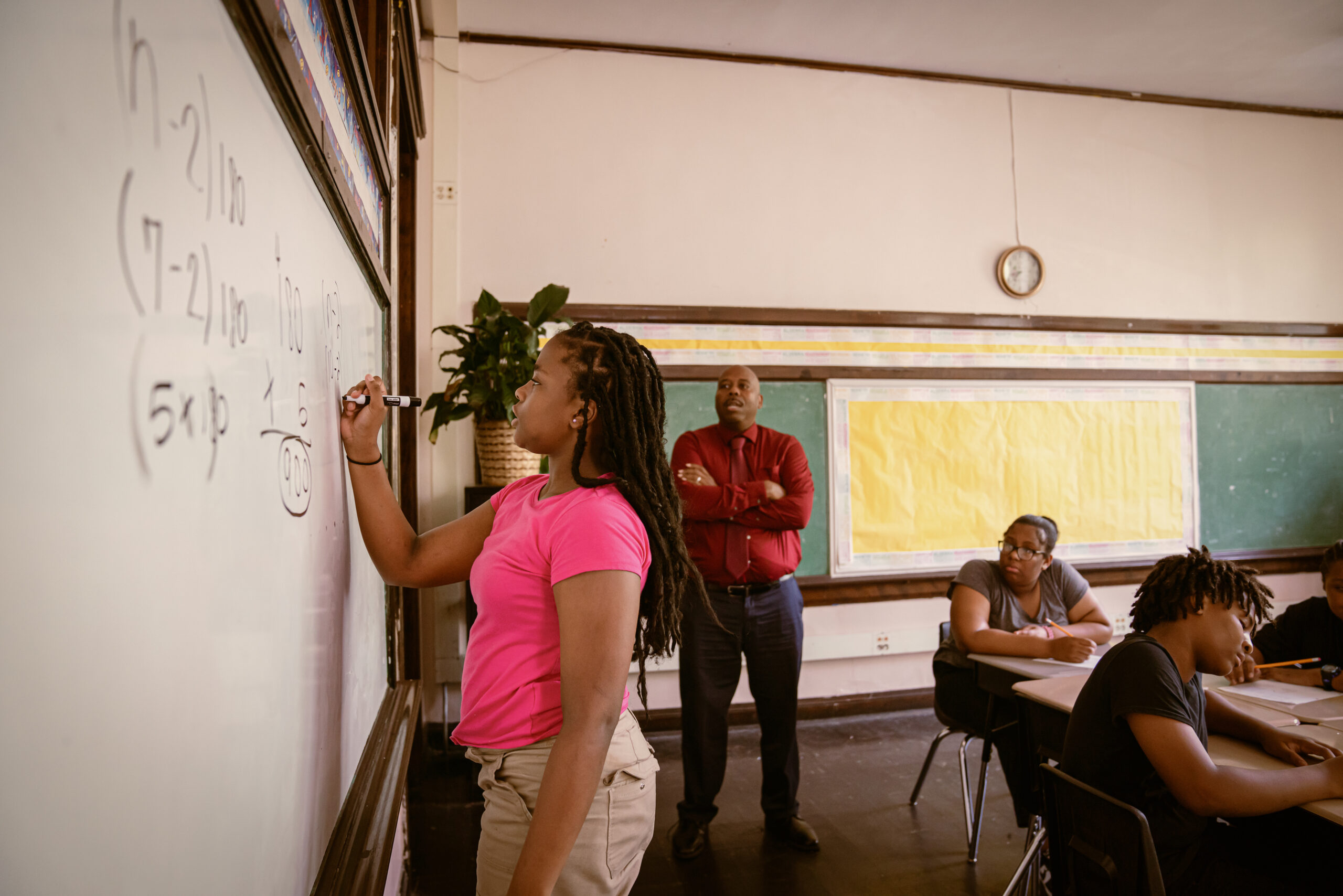 Middle school aged Black female student answering a math problem on a white board while being watched by her teacher and classmates.