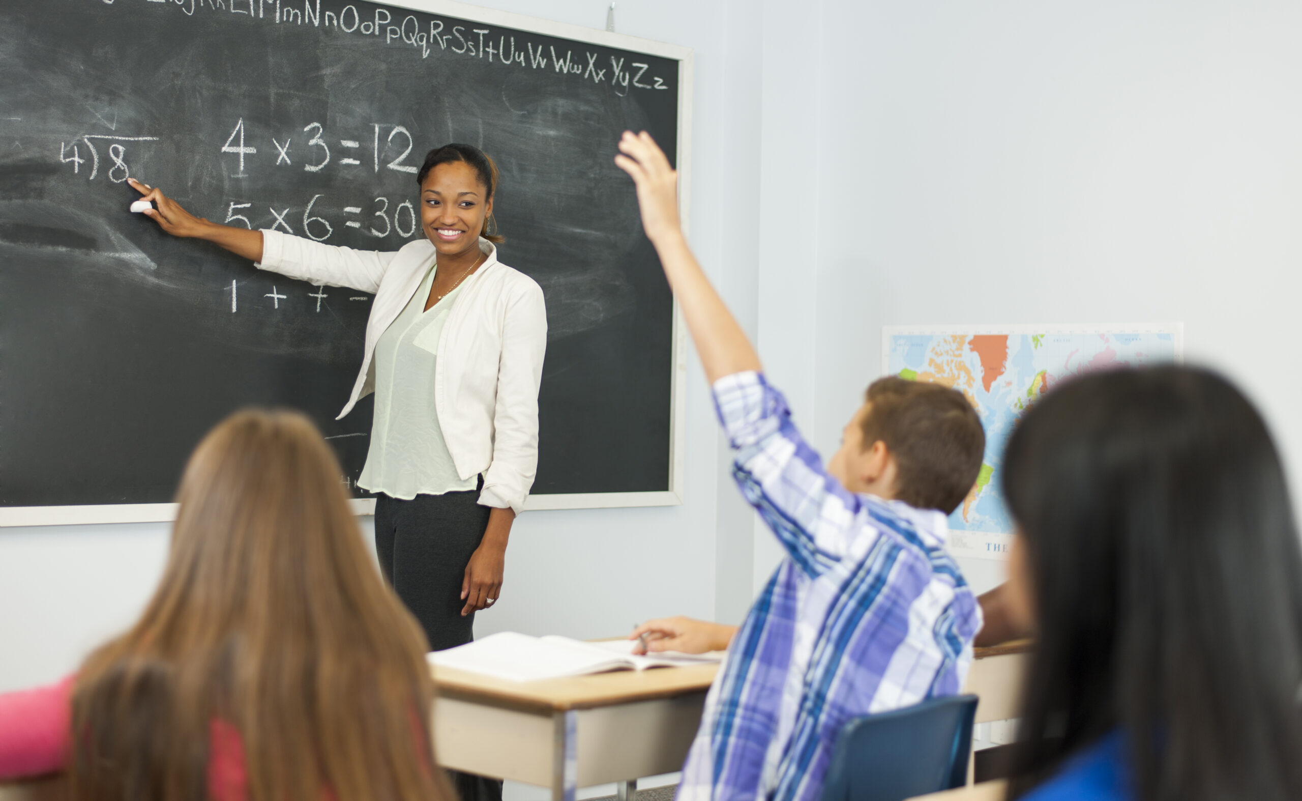 Black female teacher pointing at a division problem on chalkboard during math class with student raising their hand.