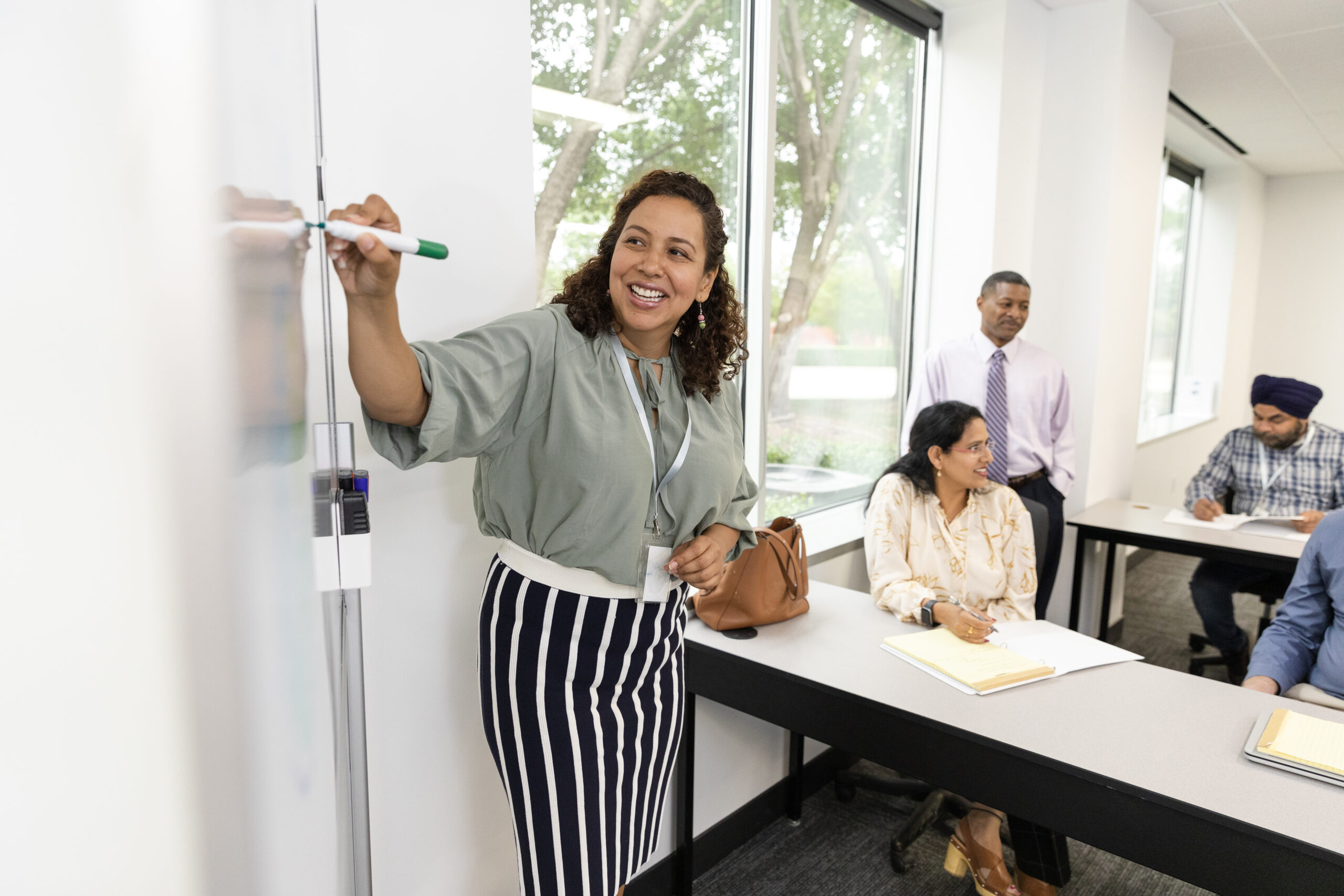 A smiling adult female writes ideas on the whiteboard as she leads curriculum professional learning for a diverse group of educators.